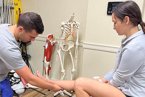 图为一名年轻男子在医务室帮助一名腿部受伤的年轻女子