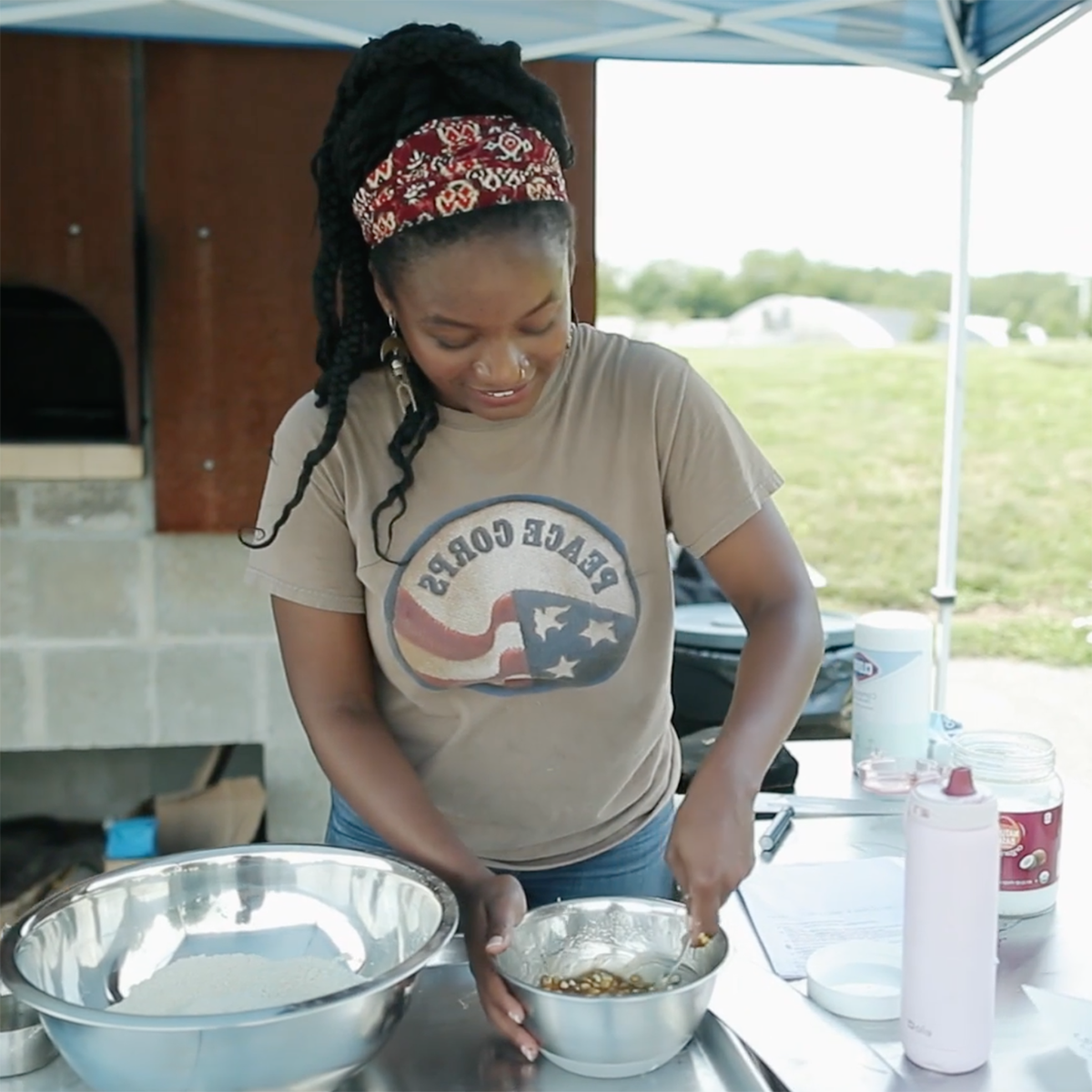 图为一位穿着和平队衬衫的年轻黑人妇女, 在伊甸园大厅校园面包烤箱里，微笑着搅拌着碗里的食物