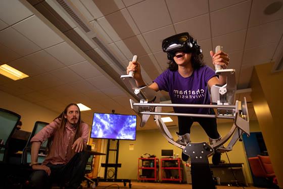 图为一名波胆网站的学生在沉浸式媒体实验室的虚拟现实机器里, 一位教授看着