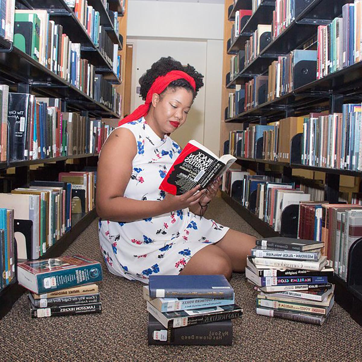 凯特琳·亨特摄, 年轻的黑人妇女, 她坐在两个图书馆书架之间，面前放着一摞书. 她正在读一本名为《黑人女作家，1950-1980》的书