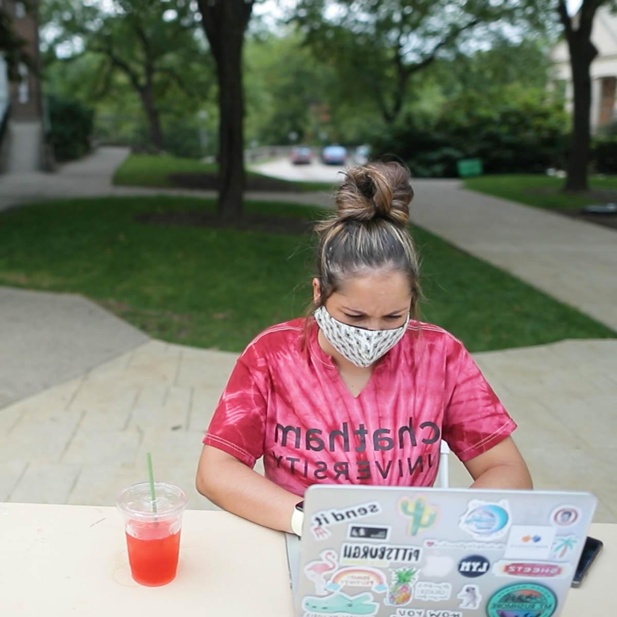 图为一名身穿扎染波胆网站衬衫的年轻女学生, 戴着口罩，坐在足球波胆平台的一张桌子旁