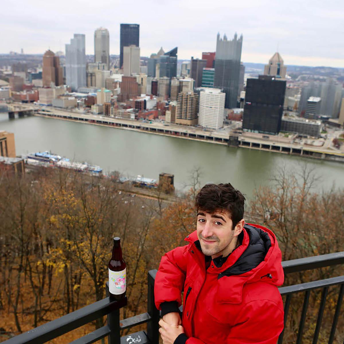 图为一名身穿红色夹克的年轻人在匹兹堡的天际线前拿着啤酒瓶摆姿势