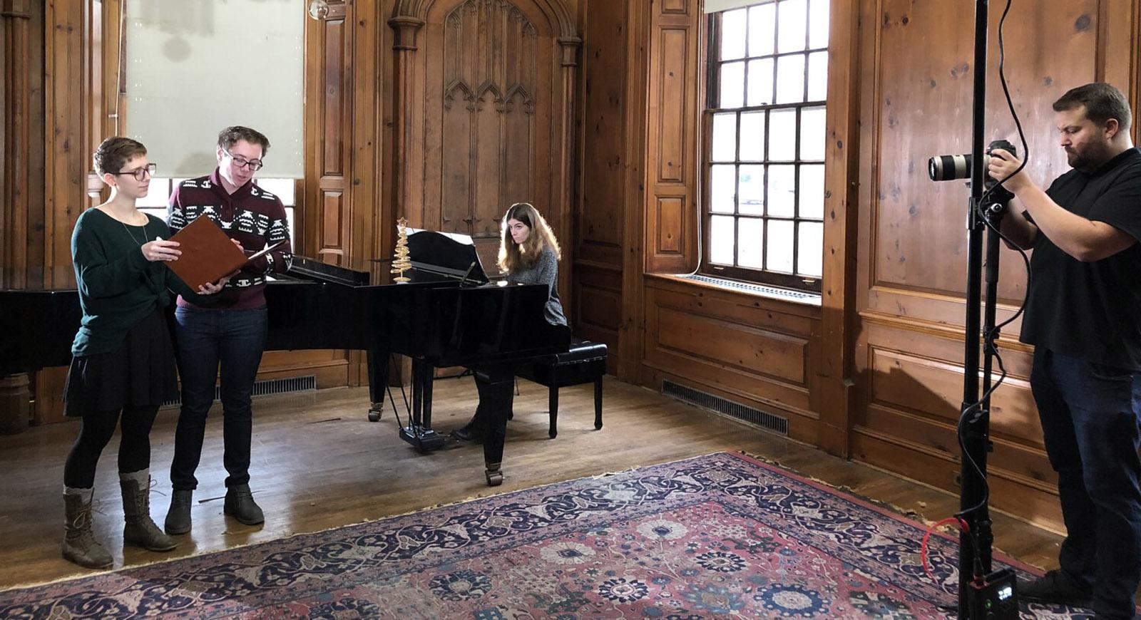 两名学生站着唱歌，一名女子在他们身后弹奏三角钢琴. 照片左边的人正在给他们拍照.